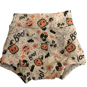 May include: White baby shorts with a Halloween-themed pattern. The design includes smiling pumpkins, skulls, spiderwebs, bats, flowers, and the word "Boo!" in black. The shorts have a ruffled edge.