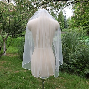 May include: A white bridal veil with a satin edge, displayed on a mannequin against a green grassy background.