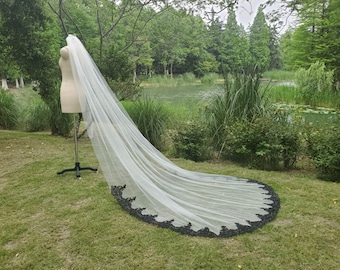 May include: A long, white tulle wedding veil with a black lace trim. The veil is draped over a mannequin and is displayed on a grassy lawn.