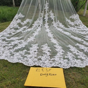 May include: A long, white bridal veil with intricate floral lace detailing along the edges. The veil is spread out on green grass, showcasing its delicate design. The text "Etsy DongXiao" is visible on a yellow card.