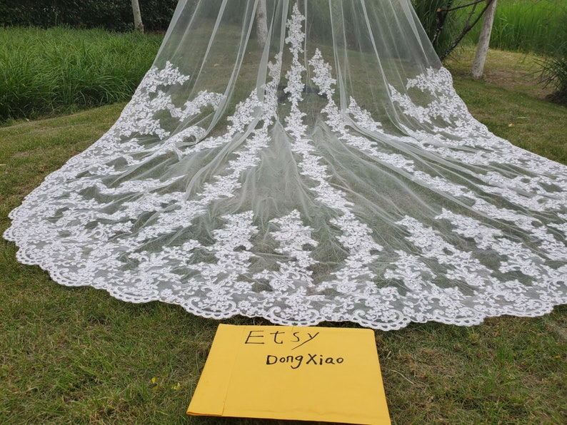 May include: A long, white bridal veil with intricate floral lace detailing along the edges. The veil is spread out on green grass, showcasing its delicate design. The text "Etsy DongXiao" is visible on a yellow card.