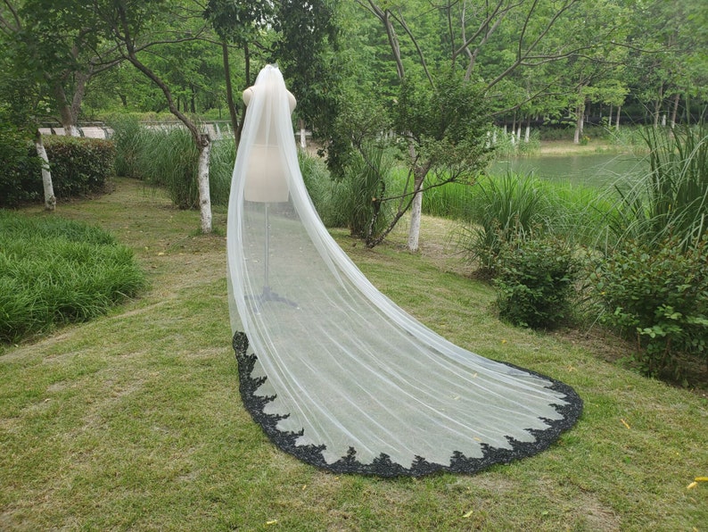 May include: A long white bridal veil with a black lace trim. The veil is flowing in the wind and is set against a backdrop of green grass and trees.