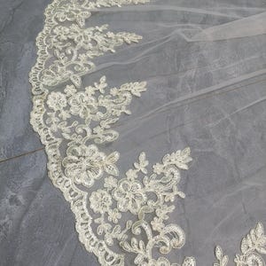 May include: Ivory-colored bridal veil with intricate floral lace detailing along the edge. The delicate lace features various flower and leaf patterns, creating a romantic and elegant design. The sheer fabric allows for a soft, flowing appearance, perfect for weddings.
