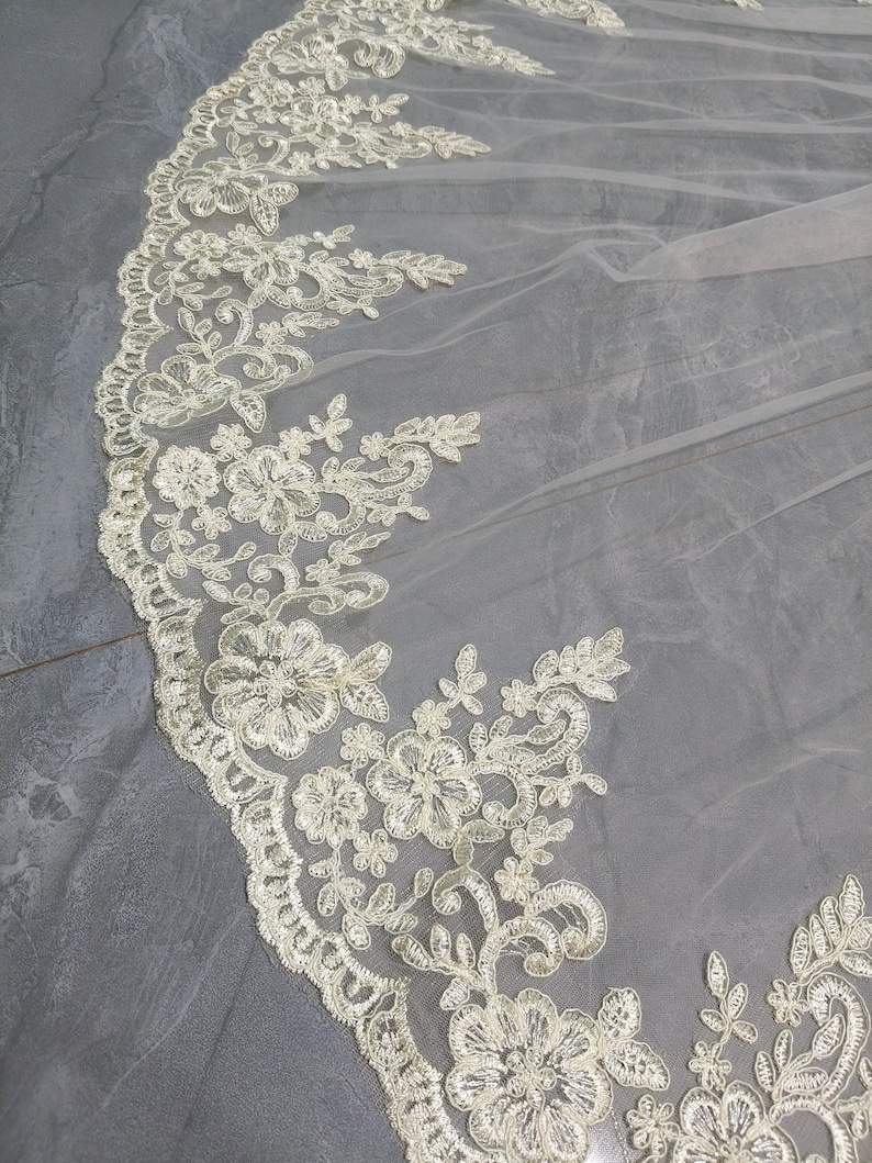 May include: Ivory-colored bridal veil with intricate floral lace detailing along the edge. The delicate lace features various flower and leaf patterns, creating a romantic and elegant design. The sheer fabric allows for a soft, flowing appearance, perfect for weddings.