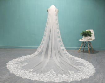 May include: A long, white bridal veil with intricate lace detailing along the edges. The veil is displayed on a mannequin, showcasing its flowing design. The background features a light blue wall and a white chair with a plant.