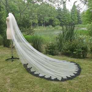 May include: A long white bridal veil with a black lace trim. The veil is draped over a mannequin and is displayed outdoors on a grassy lawn.