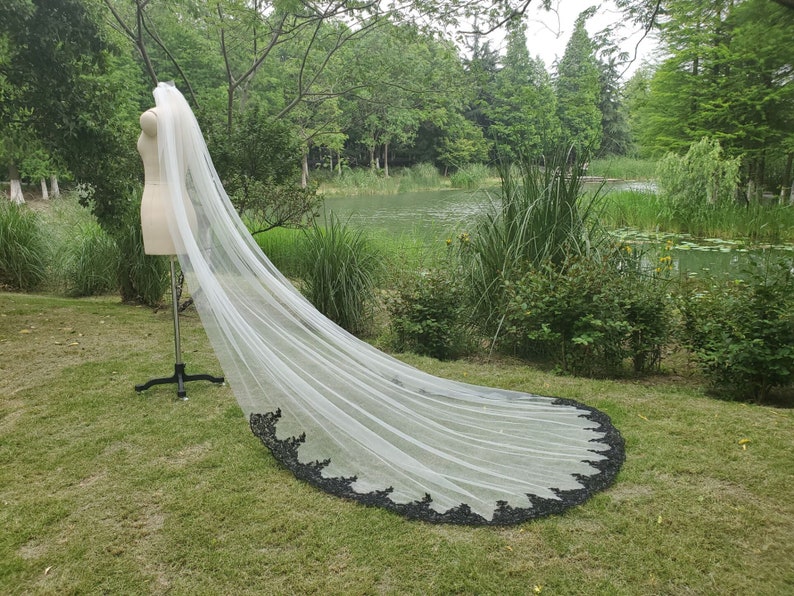 May include: A long white bridal veil with a black lace trim. The veil is draped over a mannequin and is displayed outdoors on a grassy lawn.