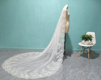 May include: A long, white bridal veil with intricate floral lace detailing along the edges. The veil is displayed on a mannequin, showcasing its flowing design. The veil's train extends across the floor, creating an elegant and romantic look.