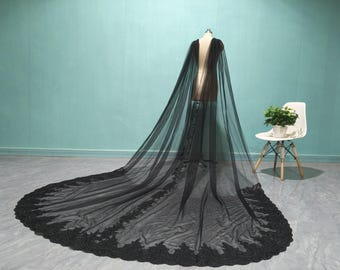 May include: A black tulle cape with a long train, displayed on a mannequin. The cape features a deep V-neck and is adorned with delicate lace detailing along the edges of the train. The background is a light teal wall and a white chair.