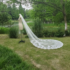 May include: A long, white bridal veil with intricate lace detailing along the edges. The veil is displayed on a mannequin in a grassy outdoor setting, with trees and a body of water in the background.