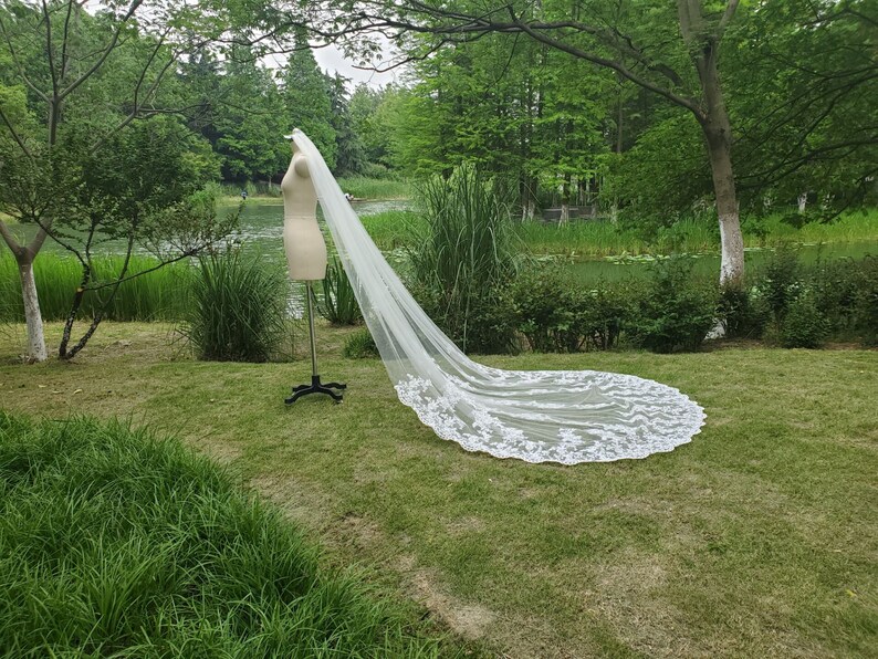 May include: A long, white bridal veil with intricate lace detailing along the edges. The veil is displayed on a mannequin in a grassy outdoor setting, with trees and a body of water in the background.