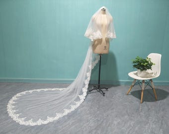 May include: A white bridal veil with a long train and delicate lace trim. The veil is displayed on a mannequin, showcasing its intricate design and flowing fabric. The background is a light blue wall.
