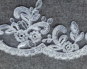 Puede incluir: Borde de encaje blanco con un diseño floral. El encaje tiene un borde festoneado y presenta delicadas flores y hojas.