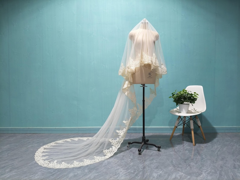 May include: A bridal veil on a mannequin. The veil is ivory with delicate lace trim along the edges. The long train extends across the floor. The mannequin is on a black stand, and a white chair with a plant sits nearby.
