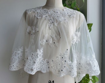 May include: White lace bridal cape with a scalloped edge and silver embellishments. The cape is on a white dress form.