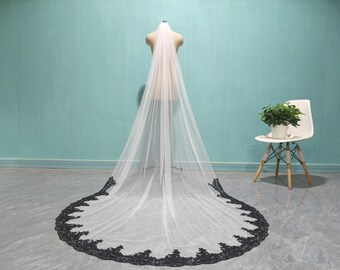 May include: A long, white bridal veil with a black floral lace trim. The veil is displayed on a mannequin, with a chair and potted plant in the background. The veil's length extends to the floor.