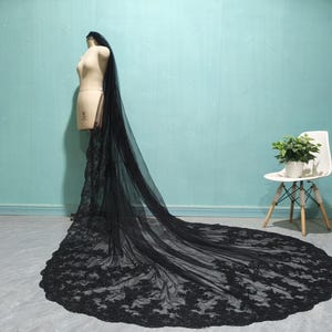 May include: A black lace wedding veil with a long train. The veil is draped over a mannequin and is displayed against a light blue background.
