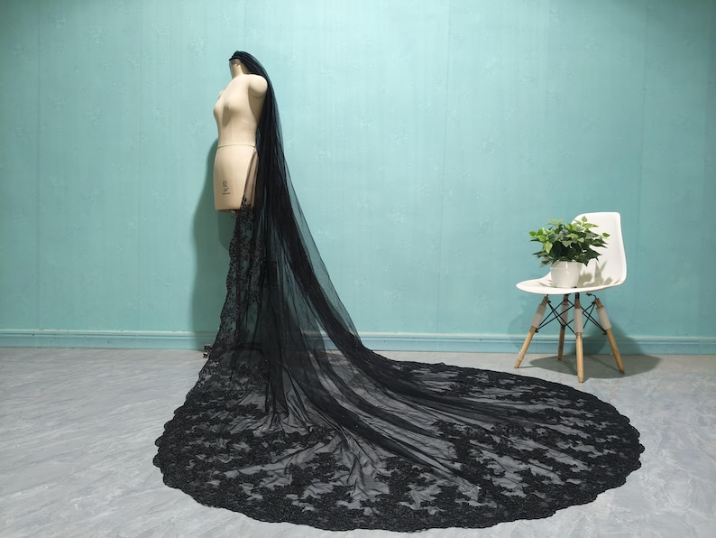 May include: A black lace wedding veil with a long train. The veil is draped over a mannequin and is displayed against a light blue background.