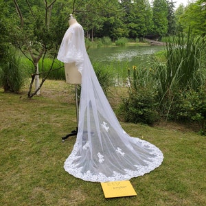 Puede incluir: Velo de novia de encaje blanco con borde festoneado y bordado floral. El velo está drapeado sobre un maniquí y se exhibe al aire libre con un fondo de hierba verde y un estanque.