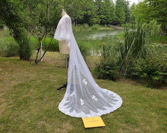 May include: White lace bridal veil with a scalloped edge and floral embroidery. The veil is draped over a mannequin and is displayed outdoors against a backdrop of green grass and a pond.