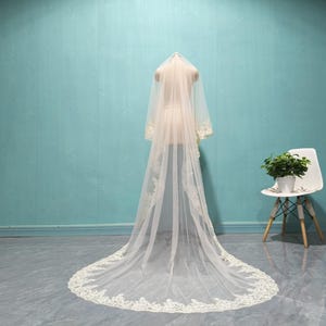 May include: A long, ivory-colored wedding veil with delicate lace trim is draped over a mannequin. The veil is displayed against a light blue wall and a gray floor. A white chair with wooden legs and a small potted plant are in the background.