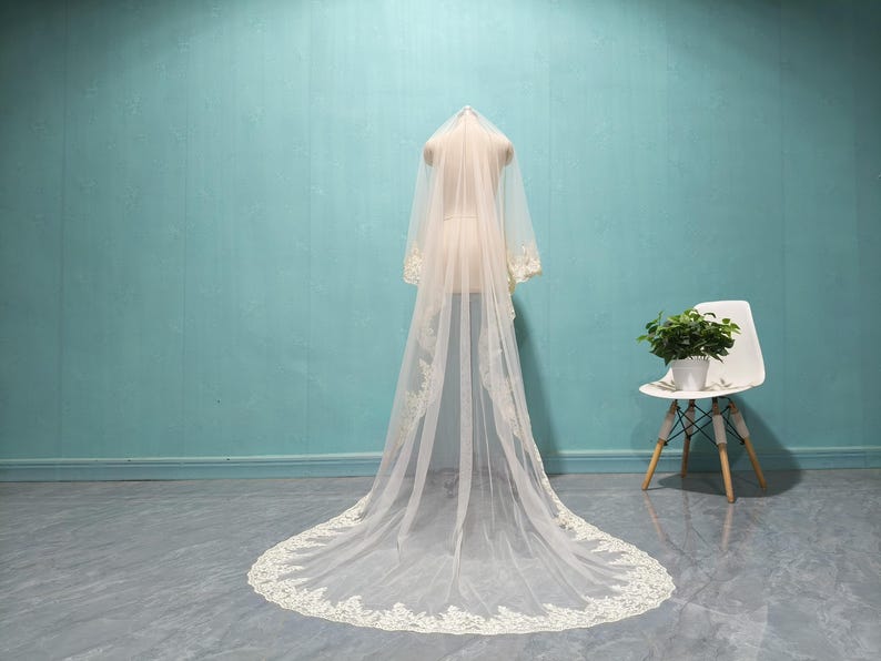 May include: A long, ivory-colored wedding veil with delicate lace trim is draped over a mannequin. The veil is displayed against a light blue wall and a gray floor. A white chair with wooden legs and a small potted plant are in the background.
