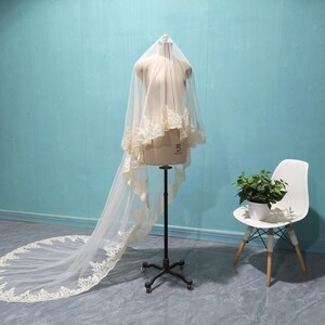 May include: A bridal veil on a mannequin. The veil is a soft, off-white color with delicate lace trim along the edges. The veil is long, with a train that extends onto the floor. The mannequin is on a black stand, and a white chair with a plant sits nearby.