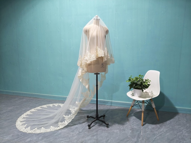 May include: A bridal veil on a mannequin. The veil is a soft, off-white color with delicate lace trim along the edges. The veil is long, with a train that extends onto the floor. The mannequin is on a black stand, and a white chair with a plant sits nearby.