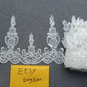 May include: White lace trim with a delicate floral pattern and sequins. The trim is folded and displayed on a gray surface. The text "Etsy DongXiao" is written on a yellow envelope.