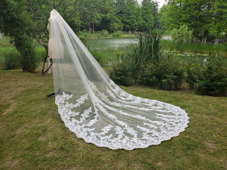 May include: A long, white bridal veil with intricate floral lace detailing along the edges. The veil is displayed on a mannequin in a grassy outdoor setting, with a lake and trees in the background. The veil's length is visible.
