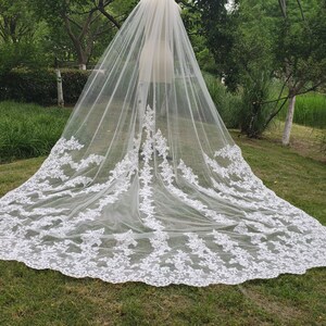 May include: A long, white bridal veil with intricate floral lace detailing along the edges. The veil is made of a sheer, flowing fabric, creating a delicate and elegant look. The veil is displayed outdoors, with green grass and trees in the background.