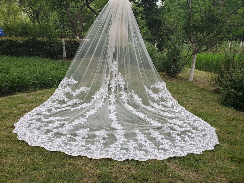 May include: A long, white bridal veil with intricate floral lace detailing along the edges. The veil is made of a sheer, flowing fabric, creating a delicate and elegant look. The veil is displayed outdoors, with green grass and trees in the background.