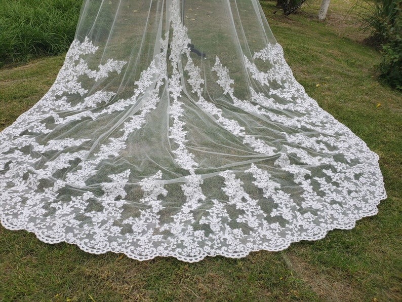 May include: A long, white bridal veil with intricate floral lace detailing along the edges. The veil is made of a sheer, lightweight fabric, and is spread out on a grassy surface. The lace design is a prominent feature.