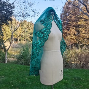 May include: A teal green sequined lace shawl draped over a dress form. The shawl has a floral design and is made of a sheer fabric.
