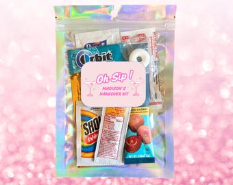 Puede incluir: Una bolsa transparente holográfica con cremallera llena de artículos para la resaca. La bolsa contiene una etiqueta rosa que dice "Oh Sip! MADISON'S HANGOVER KIT" con dos copas de martini. También contiene chicles, mentas y caramelos de cereza.