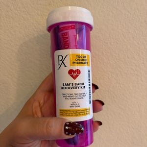 以下が含まれることがあります： 白いキャップが付いた半透明のピンク色のプラスチック製薬瓶で、中に絆創膏が入っています。ラベルには「TOO LIT OH SHIT! PHARMACY, SAM'S BACH RECOVERY KIT, DIRECTIONS: TAKE 1 AFTER A WILD NIGHT OUT TO HELP YOU BOUNCE BACK, QTY: 1, REFILLS: 0, RX#: IDGAF」と記載されています。