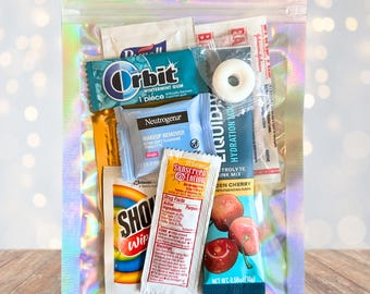 May include: A clear, iridescent zip-lock bag containing assorted small packets. Items include Orbit wintermint gum, Neutrogena makeup remover wipes, sunscreen lotion, and a cherry-flavored electrolyte drink mix. A white mint is also visible.