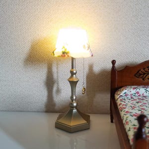 Puede incluir: Una lámpara de mesa en miniatura con una pantalla con estampado floral. La lámpara tiene una base y un tallo dorados, con una cadena de tracción. La lámpara está junto a una cama en miniatura con una colcha con estampado floral.