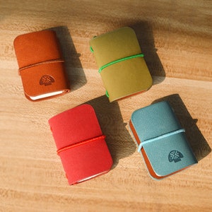 May include: Four small leather notebooks in different colors: brown, green, red, and blue. Each notebook has a string closure and a small embossed design on the front.