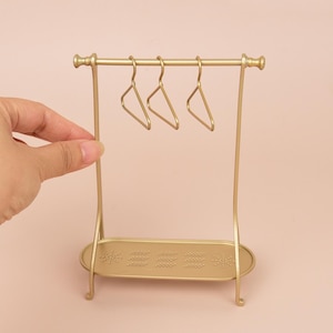 May include: A miniature gold-colored metal clothing rack featuring a horizontal bar with three matching small hangers suspended from it and an oval-shaped base tray with a decorative embossed pattern of small dots arranged in geometric clusters.