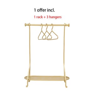 May include: A gold-colored metal clothing rack with a horizontal bar, a bottom shelf, and three matching gold-colored hangers. The text above the rack reads: 1 offer incl. 1 rack + 3 hangers. The structure features curved side supports.