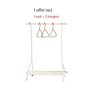 May include: A white metal clothing rack with a bottom shelf and three gold-toned triangular hangers. The text above the rack reads: 1 offer incl. 1 rack + 3 hangers. The rack features a horizontal bar supported by two curved side legs.