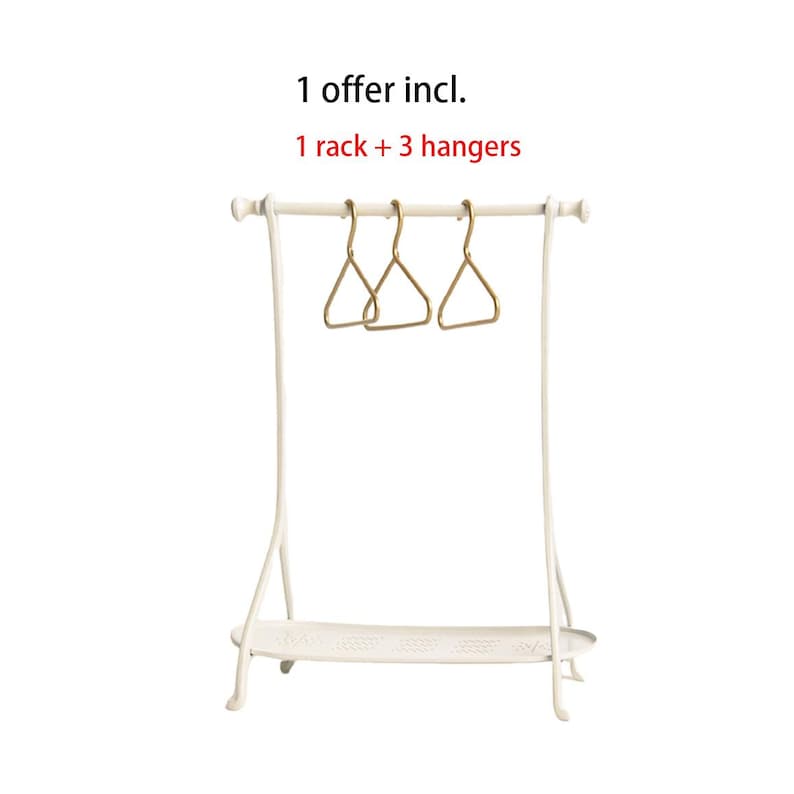May include: A white metal clothing rack with a bottom shelf and three gold-toned triangular hangers. The text above the rack reads: 1 offer incl. 1 rack + 3 hangers. The rack features a horizontal bar supported by two curved side legs.