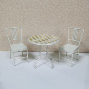 May include: A miniature white metal patio set with a round table and two matching chairs. The table has a yellow floral pattern on the tabletop. The chairs have vertical back slats.