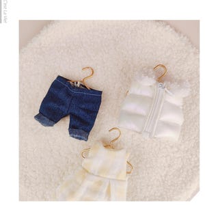 May include: Three miniature garments on gold-toned hangers: a pair of dark blue denim shorts with cuffed hems, a white puffer vest with a front zipper and fuzzy collar, and a yellow and white checkered sleeveless dress with a pleated skirt.