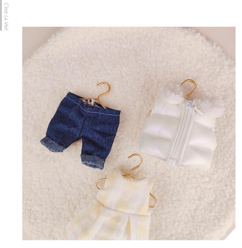 May include: Three miniature garments on gold-toned hangers: a pair of dark blue denim shorts with cuffed hems, a white puffer vest with a front zipper and fuzzy collar, and a yellow and white checkered sleeveless dress with a pleated skirt.