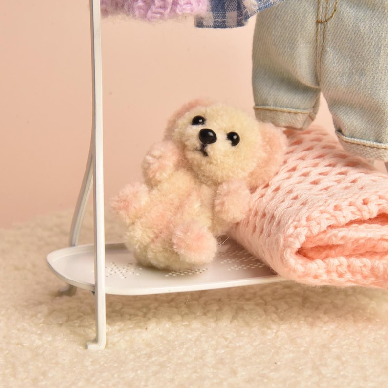 May include: A small, fuzzy, cream-colored teddy bear with black bead eyes and a black nose sits on a white metal shelf. Beside the bear is a folded light pink knit blanket, and above it hang light blue and white checkered fabric and lavender knit.