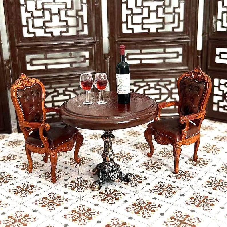 May include: A miniature dollhouse furniture set with two brown leather chairs, a round wooden table, and a bottle of wine. The chairs have ornate carvings and the table has a pedestal base. The set is on a tiled floor.