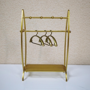 May include: A gold-colored metal garment rack featuring two horizontal bars, with the top bar accented by four small spherical details. Three matching gold-colored metal hangers are suspended from the lower bar, and a slatted shelf sits at the base.