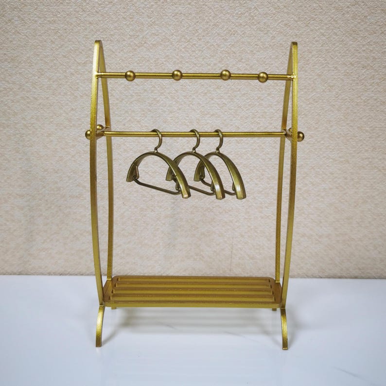 May include: A gold-colored metal garment rack featuring two horizontal bars, with the top bar accented by four small spherical details. Three matching gold-colored metal hangers are suspended from the lower bar, and a slatted shelf sits at the base.